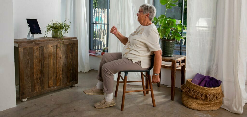 En eldre dame sitter på en stol i stua mens hun beveger på armer og ben. Foran seg på en sjenk har hun Berntsen Beinlaus, en ipad som hjelper henne med treningen.
