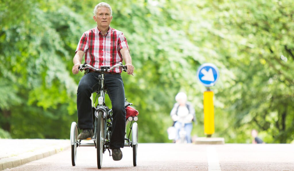 Eldre mann iført t-skjorte og bukse sykler på veien på en tradisjonell trehjulssykkel.
