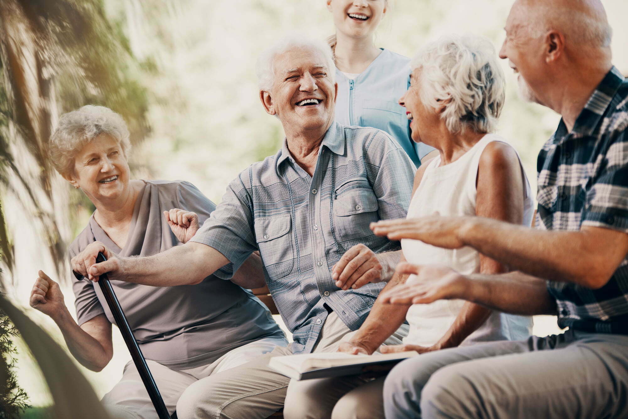 Fire smilende eldre mennesker ser på hverandre, ler og koser seg på en benk ute. En sykepleier står i bakgrunn og ser på de.