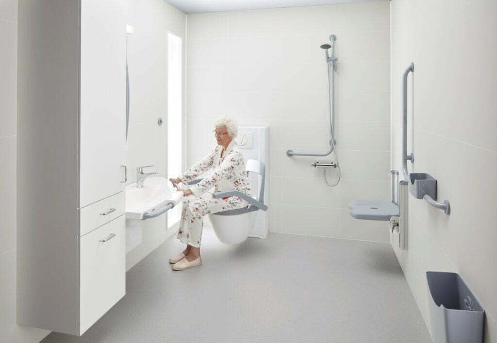 Eldre dame sitter i pysjen inne på et baderom på et BVT toalett. Toalettet er vridd mot vasken, og damen holder seg i håndtakene rundt vasken. Hun smiler mens hun ser på vasken.