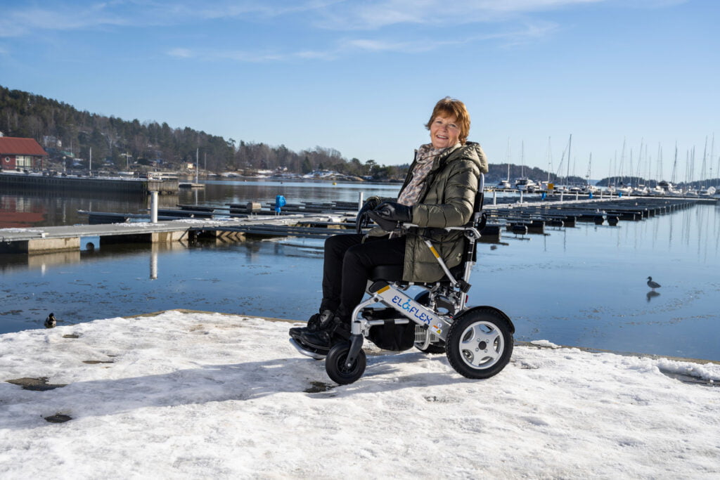 Berit sitter i den elektriske rullestolen Eloflex på brygga i Son. Hun smiler til kameraet. i bakgrunnen ser du sjøen og båthavna.