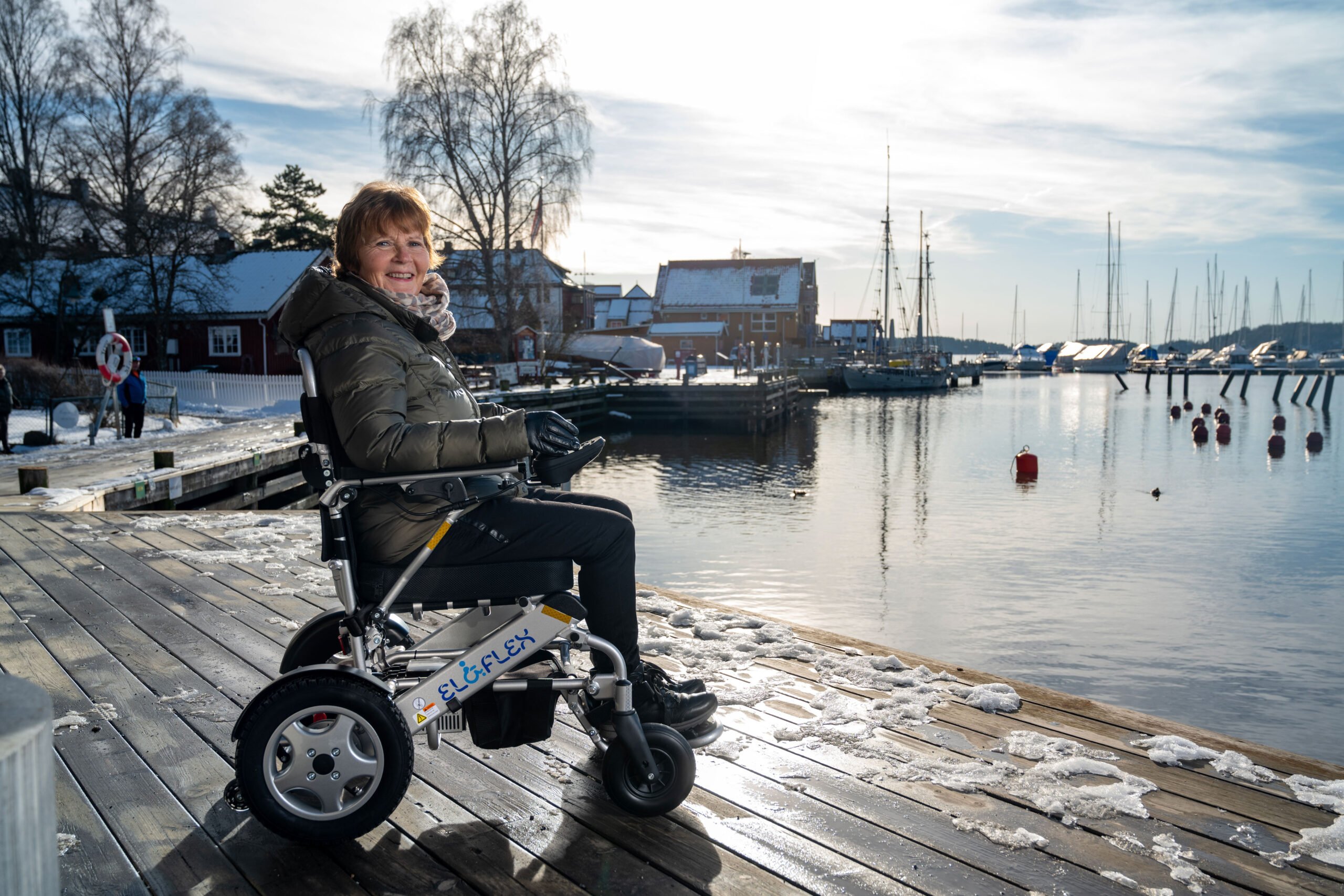 Berit sitter i rullestolen Eloflex på brygga i Son. Hun smiler til kameraet. Det er snø på brygga og hun har på seg godt med klær. Du ser sjøen, noen seilbåter og hus og restuaranter som ligger på brygga.