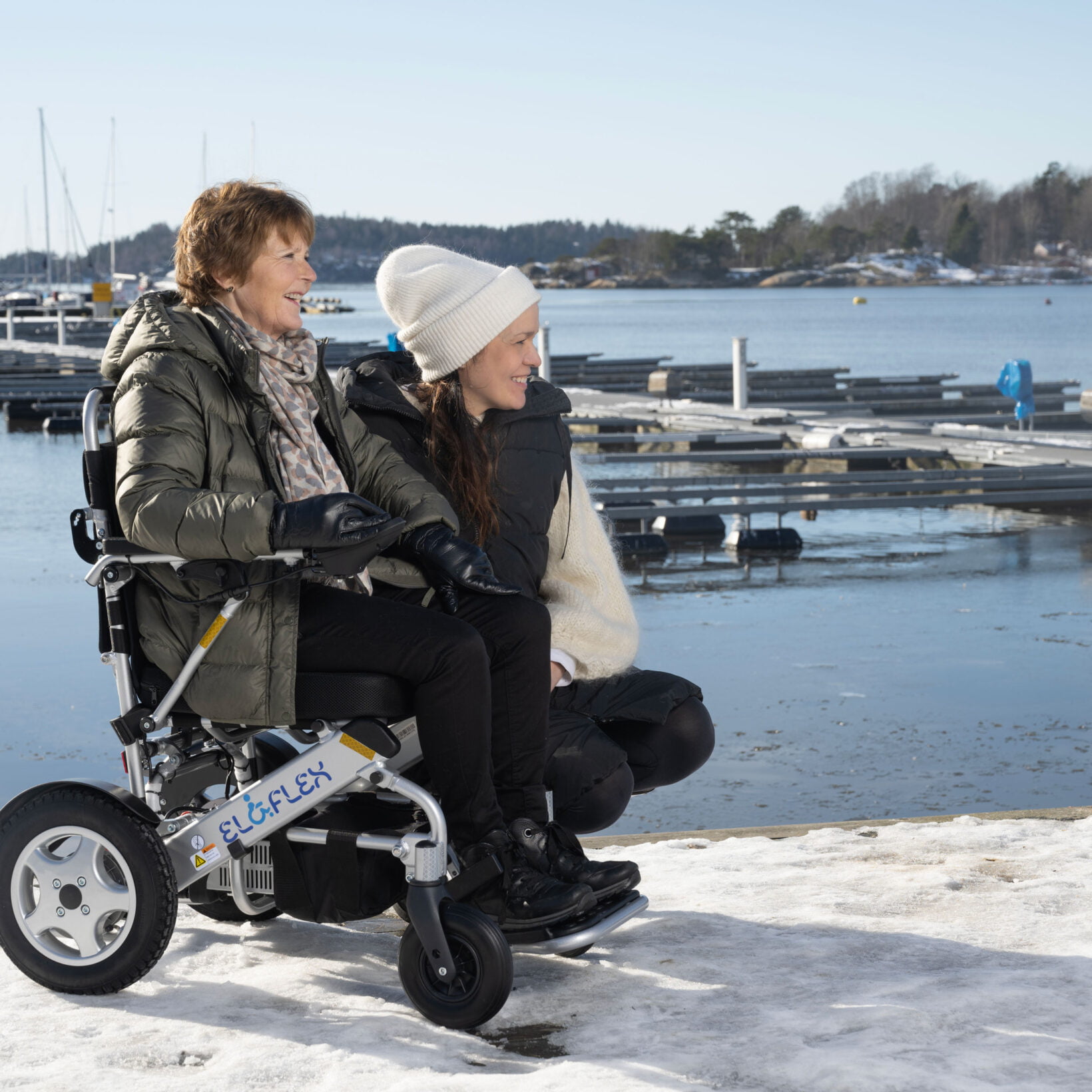Berit og Aleksandra er på brygga i Son. Berit sitter i en elektrisk rullestol. Aleksandra har satt seg ned på huk ved siden av Berit. De ser bortover på noe. Det er snø på brygga, og du ser båthavnen i bakgrunnen.