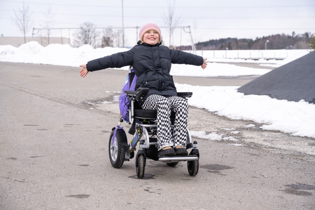 Ida stråler og smiler mens hun holder begge armene ut til siden. Hun sitter i en Eloflex elektrisk rullestol. Skolesekken har hun festet på baksiden av rullestolen.