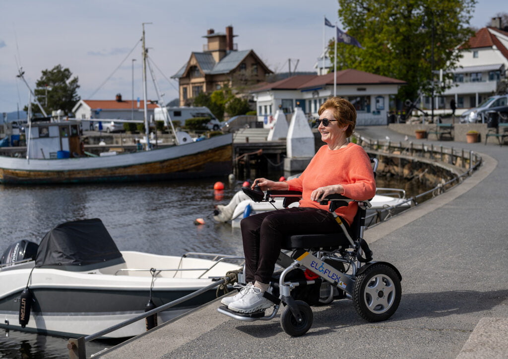 Berit sitter i rullestolen på brygga i Drøbak. Solbrillene er på og hun nyter det vårlige været. Bak henne ser du noen hus, og noen båter.