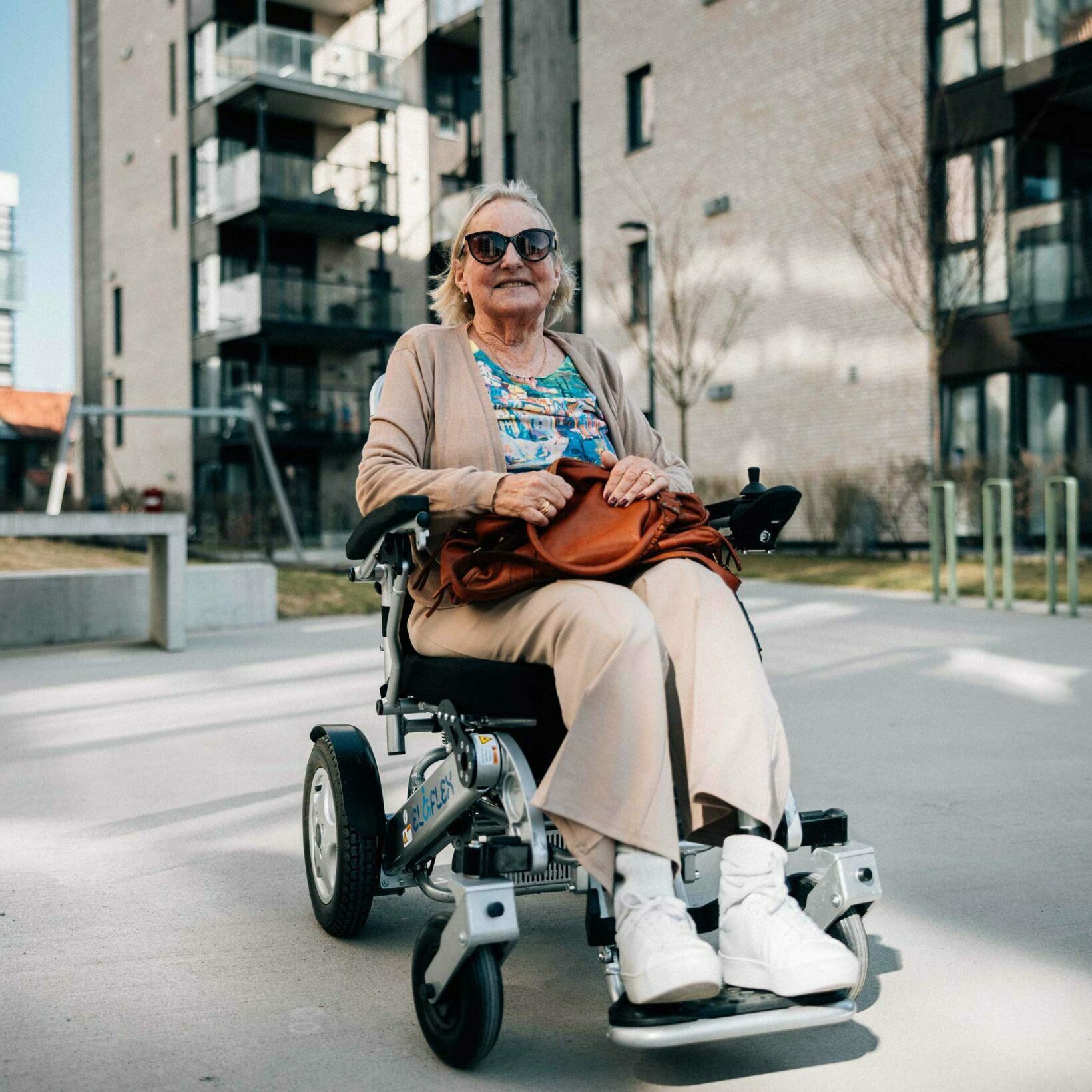 Synnøve kjører i sin Eloflex elektriske rullestol med solbrillene på og veska i fanget. Hun kjører i et boligområde med blokker.