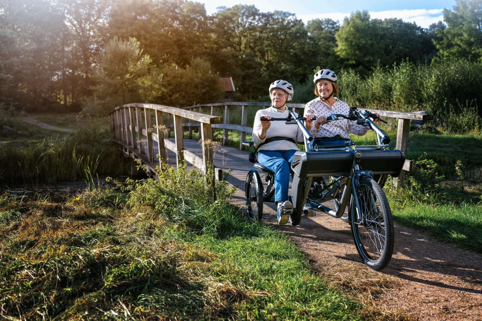 To voksne damer sykler på en side-by-side Orthros tandemsykkel. De har akkurat syklet over en bro, og er omgitt av skog og natur.