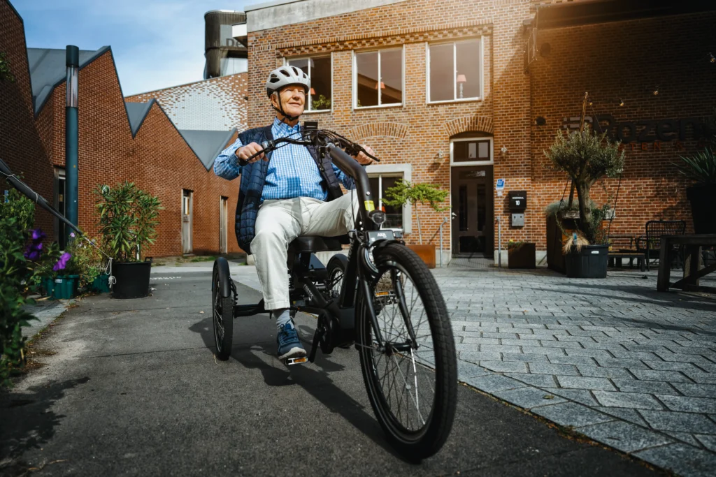 Eldre voksen mann sykler på en Cortes trehjulssykkel. Han er fornøyd, mens han sykler i en hyggelig bakgård med brostein og asfalt.