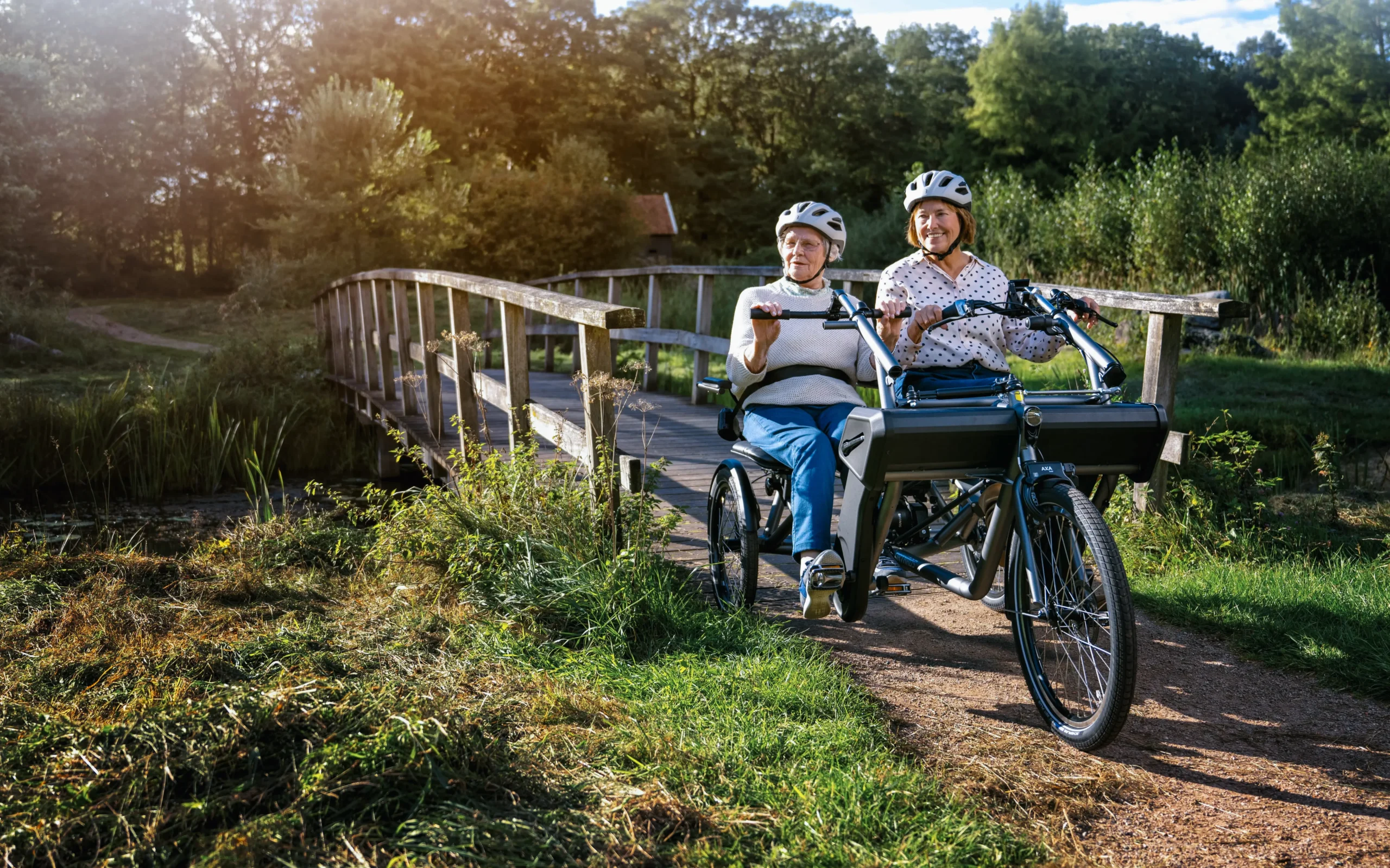 To voksne damer sykler på en side-by-side Orthros tandemsykkel. De har akkurat syklet over en bro, og er omgitt av skog og natur.