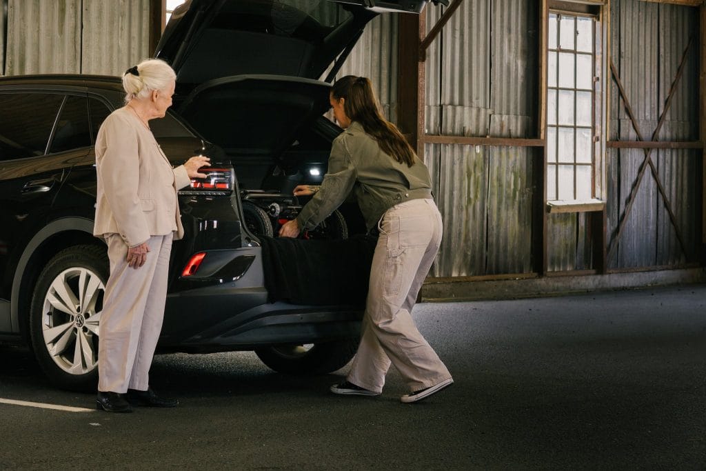 Amanda står bak bilen, og er i ferd med å løfte ut elio sammenleggbare elektriske rullestol. Laila, holder i bilen mens hun venter.