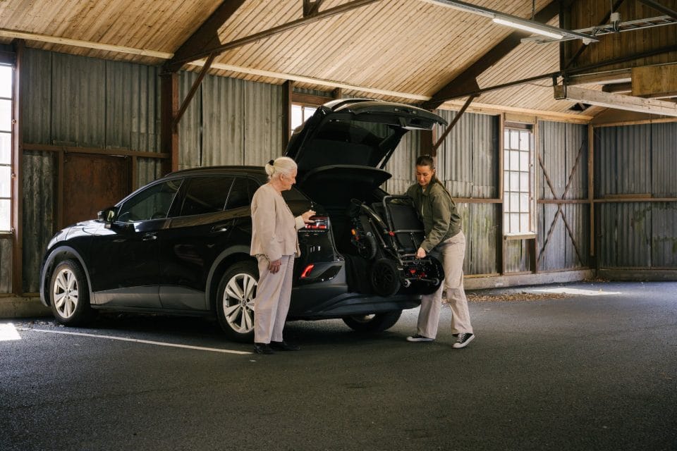 Amanda står bak bilen og løfter ut elio sammenleggbare elektriske rullestol. Bagasjeluka på bilen er åpen, og Laila som skal sitte i stilen står og ser på.