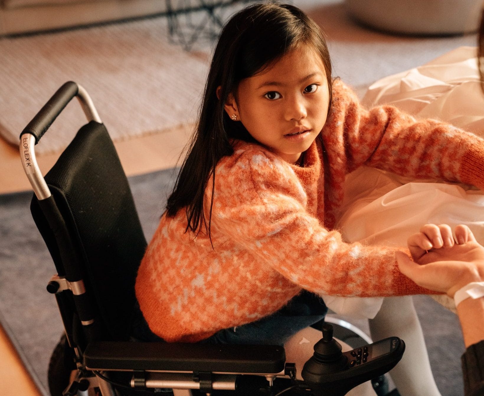 Jente sitter i en elektrisk rullestol ved et bord. Ser på kamera.