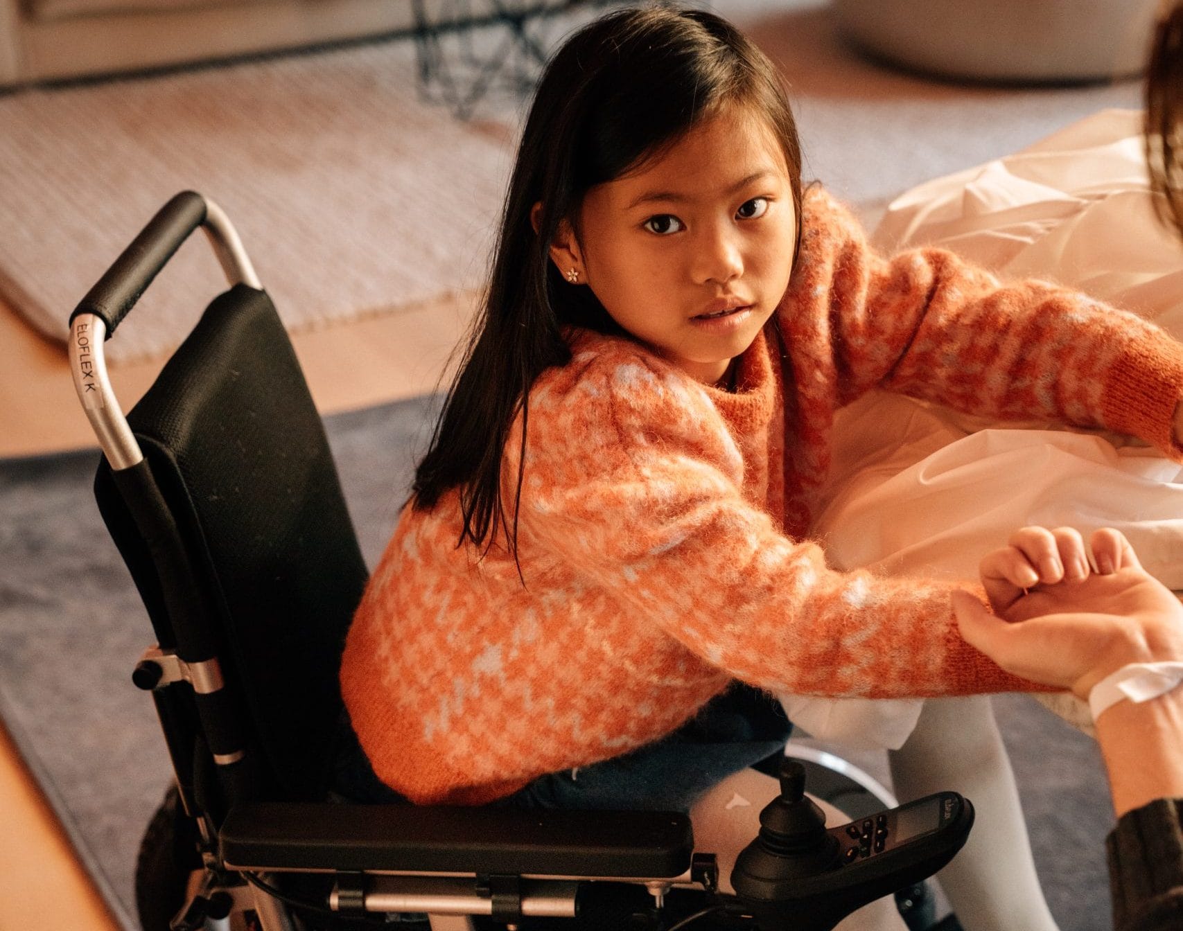 Jente sitter i en elektrisk rullestol ved et bord. Ser på kamera.