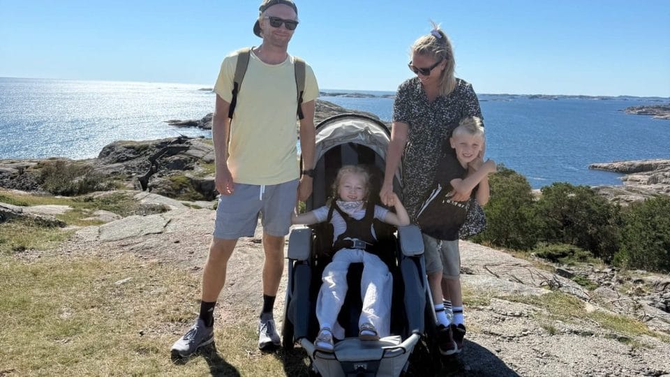 Familie på tur i solskinn. På toppen av en liten fjellknaus, med utsikt bak dem utover havet. Eldste datteren sitter i en Josi Wismi E-drive.