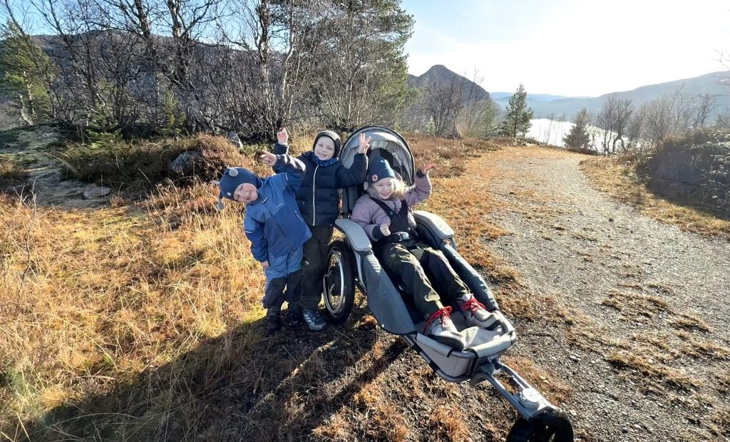 Søsken på skogstur. Eldste jenta sitter i en Josi Wismi E-drive. De vinker til kameraet. I bakgrunnen er det fjell og vann.