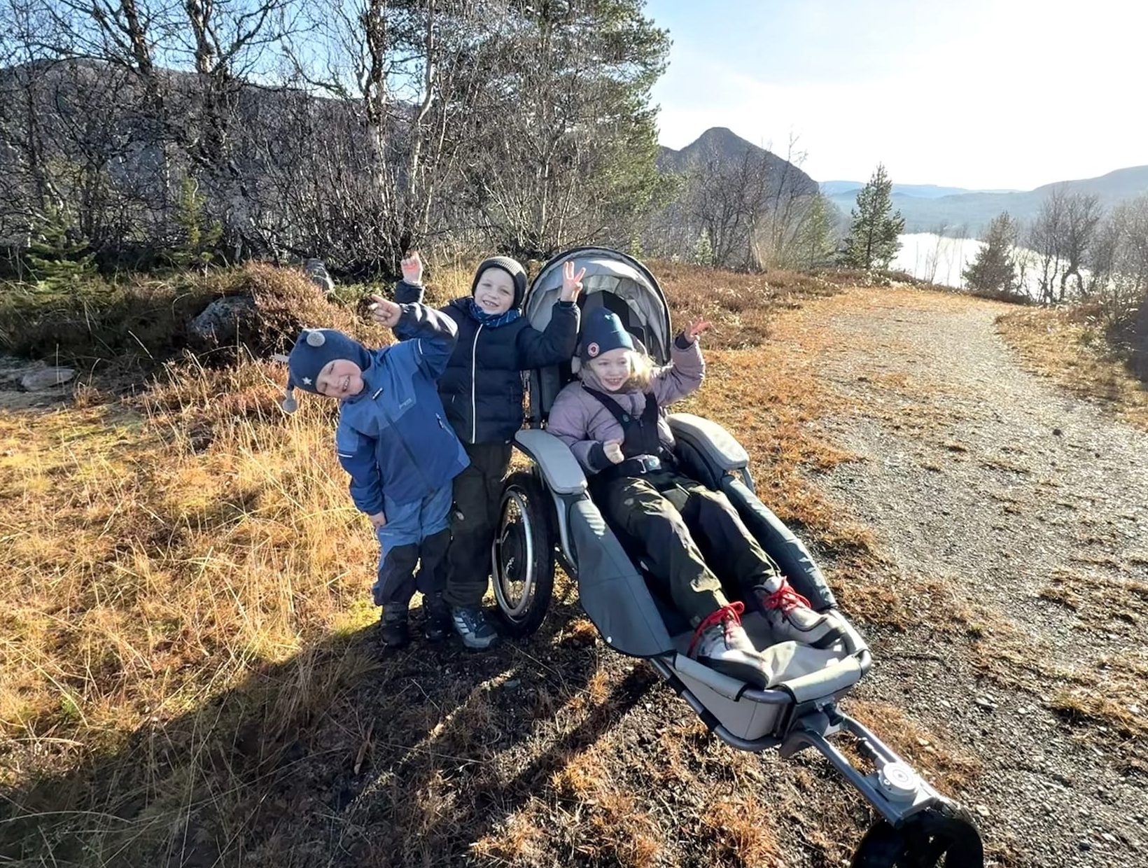 Søsken på skogstur. Eldste jenta sitter i en Josi Wismi E-drive. De vinker til kameraet. I bakgrunnen er det fjell og vann.