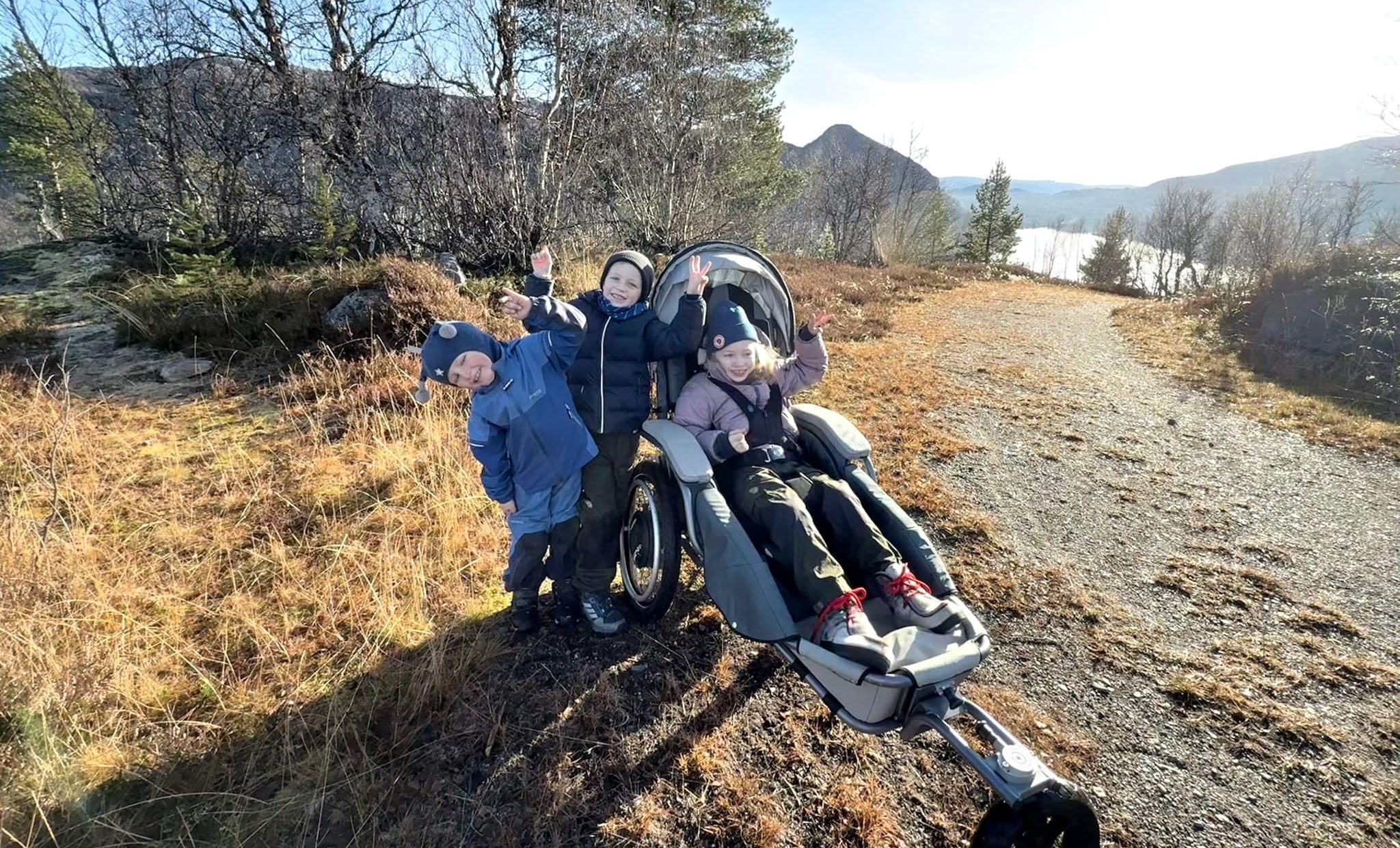Søsken på skogstur. Eldste jenta sitter i en Josi Wismi E-drive. De vinker til kameraet. I bakgrunnen er det fjell og vann.