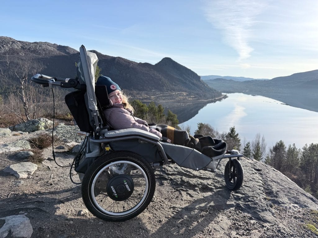 Ella smiler og sitter godt påkledd i vognen Josi Wismi E-drive. Vognen Står på en fjelltopp med utsikt over vann og fjell.