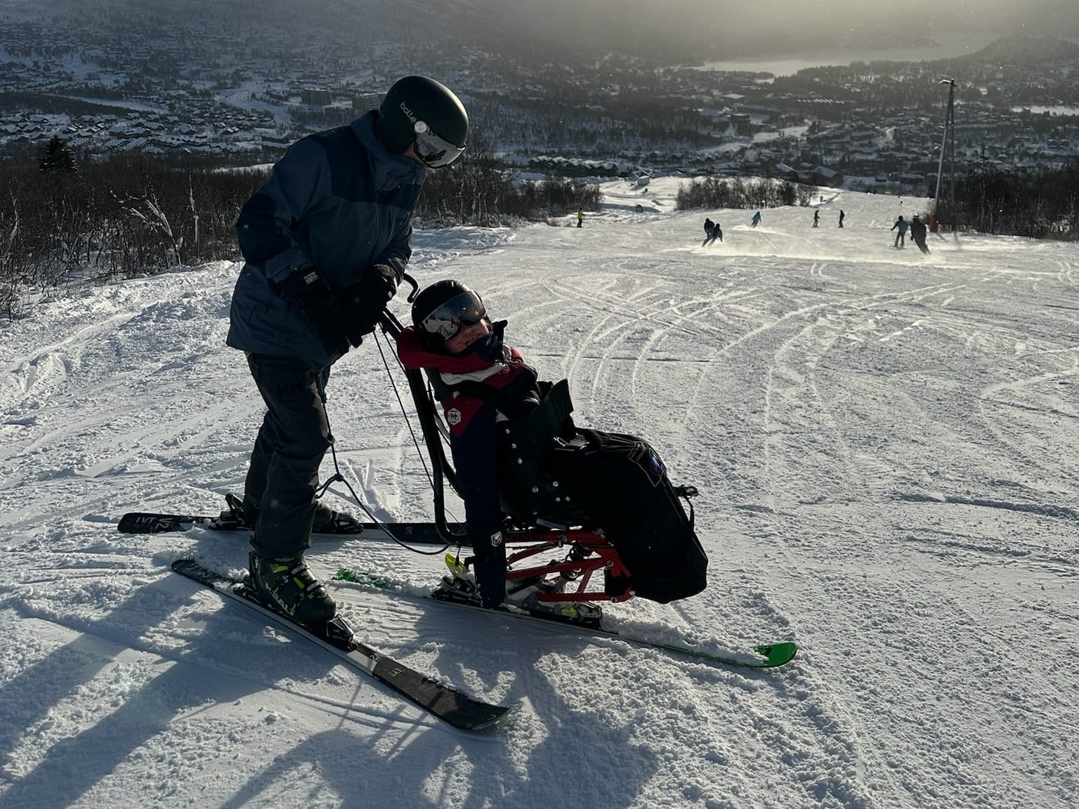 Far og datter kjører sitski i slalåmbakken. Sola skinner, og utsikten ligger bak dem.