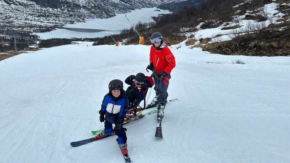 Pappa, lillebror og Ella i slalåmbakken. De smiler og ser på kamera. Ella kjører sitski.