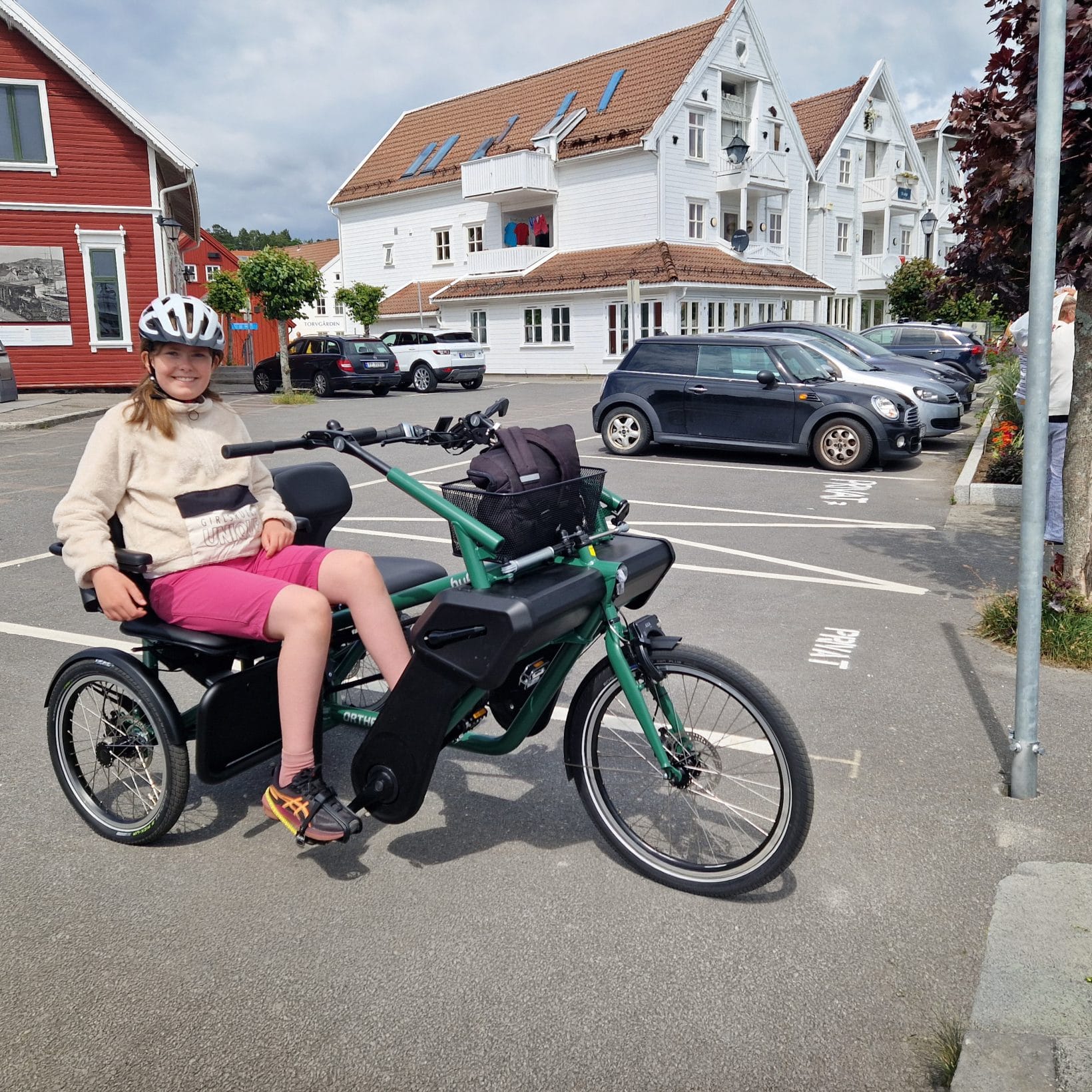 En jente på 11 år sitter på Orthros tandemsykkel på en parkeringsplass på sørlandet. Hun har på hjel, lys genser og shorts.