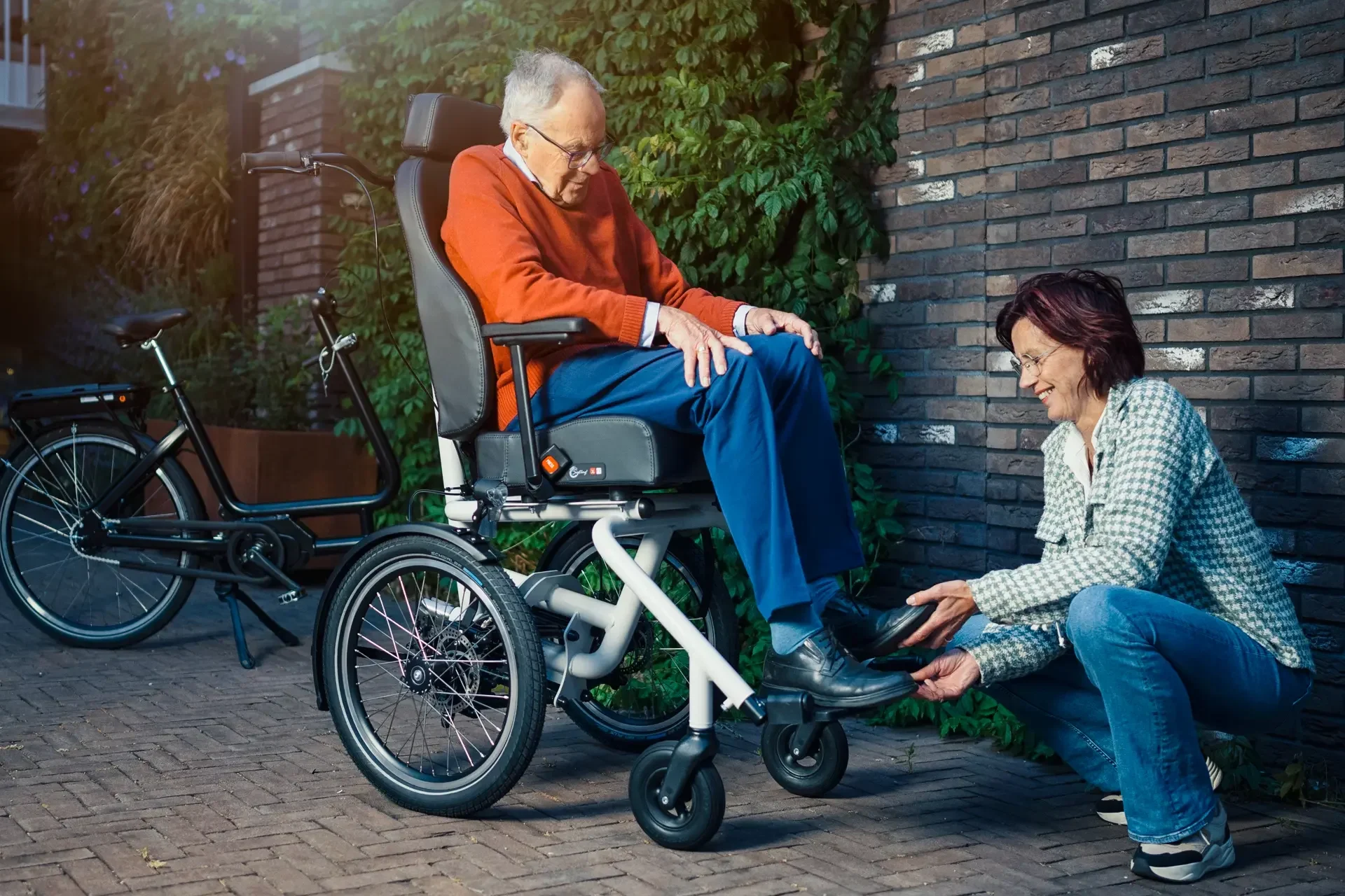 Eldre mann sitter foran i tandemsykkelen Diaz. En voksen dame står på huk, mens hun hjelper mannen med å ta på skoa.