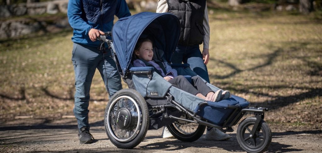 Jente som smiler, de er på tur, og hun sitter i den elektriske terrengvogna Josi Wismi E-drive G2. Foreldrene står bak, de ser du bare deler av. De er på trilletur med en gressbakke ved siden av seg.