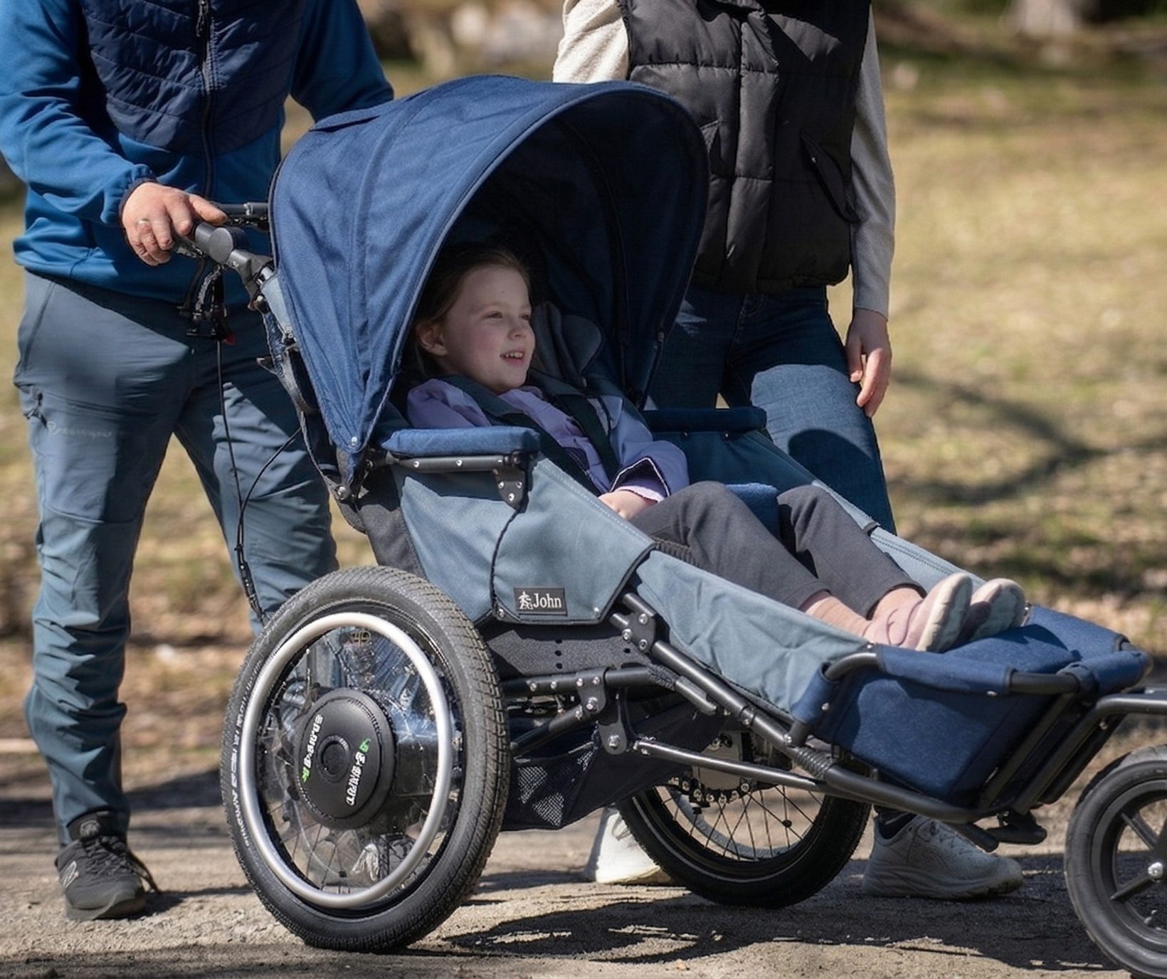 Jente som smiler, de er på tur, og hun sitter i den elektriske terrengvogna Josi Wismi E-drive G2. Foreldrene står bak, de ser du bare deler av. De er på trilletur med en gressbakke ved siden av seg.