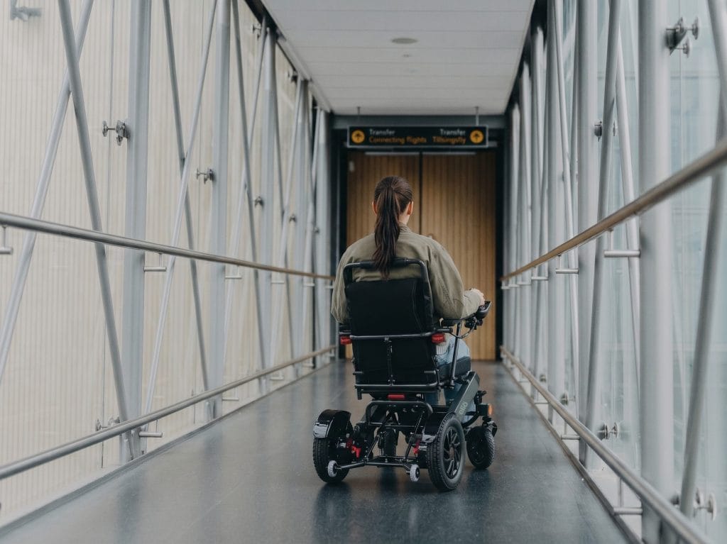 Dame sitter i elio elektriske rullestol på vei inn på et fly. Du ser henne bakfra i broa over til flyet.