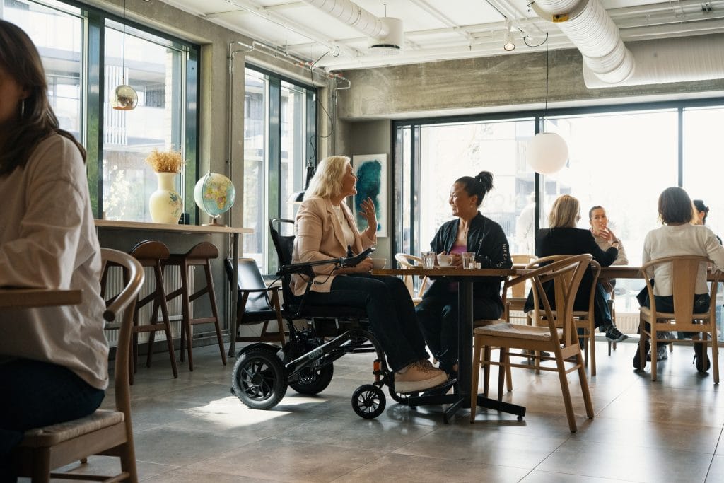 En pent kledd dame sitter i Elio sammenleggbare elektriske rullestol på Café med en venninne. De ser på hverandre og prater. Rundt dem ser du andre gjester.