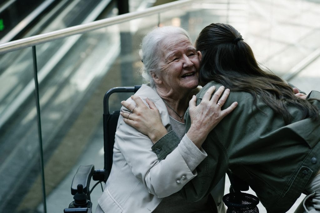 Bestemor og barnebarn klemmer. Barebaret står med ryggen til, og bestemoren smiler. Bestemor sitter i elio sammenleggbare elektriske rullestol.
