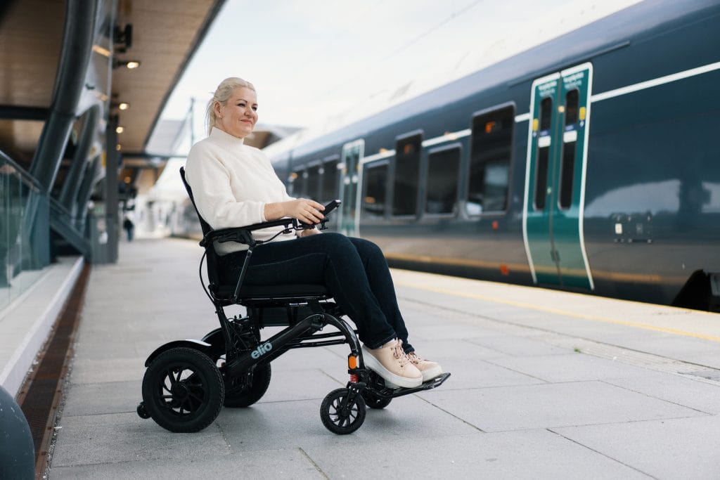 Marianne sitter i Elio elektrisk rullestol og venter på toget. Hun er iført hvit genser og mørk bukse. Toget står på perrongen.