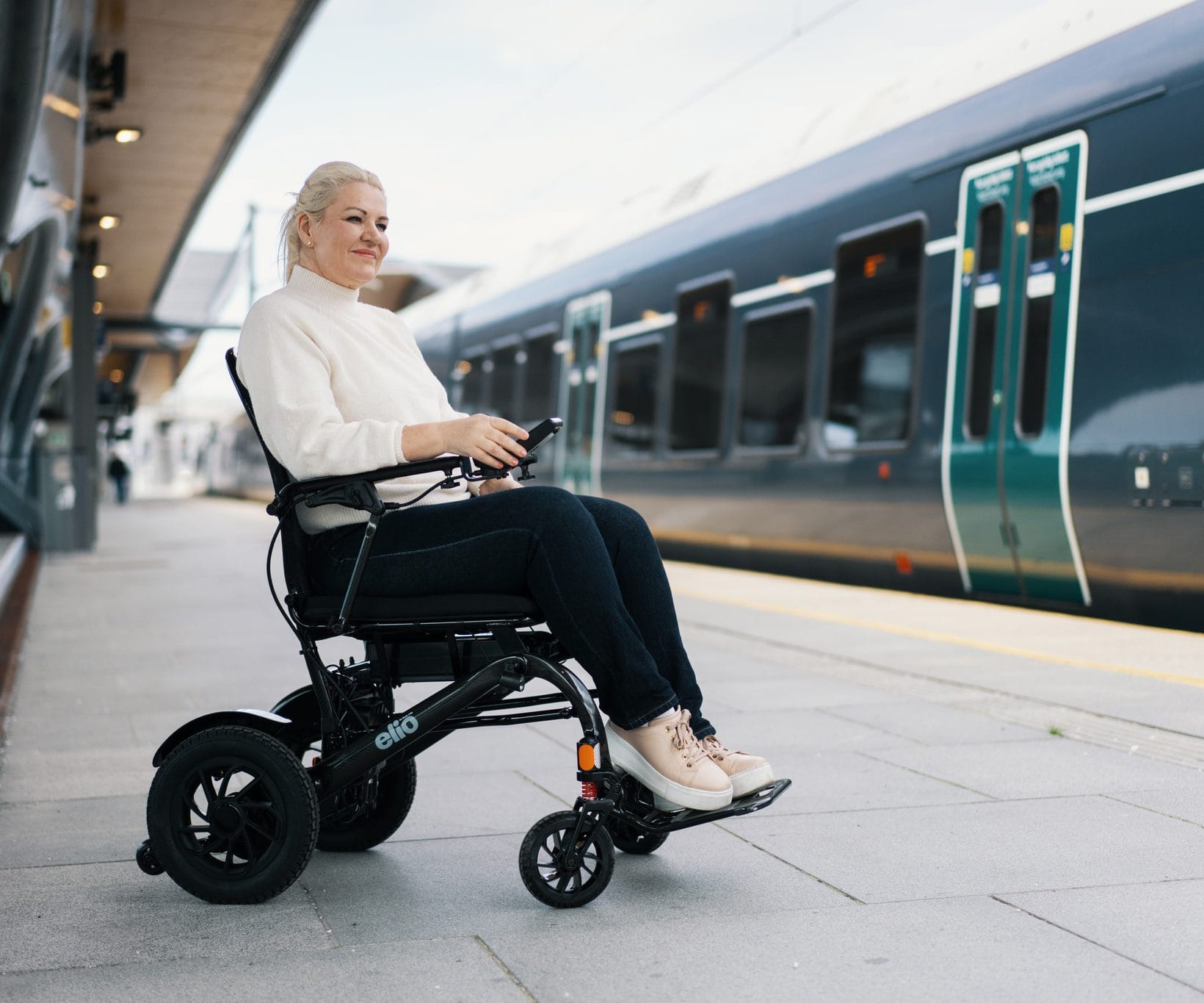 Marianne sitter i Elio elektrisk rullestol og venter på toget. Hun er iført hvit genser og mørk bukse. Toget står på perrongen.