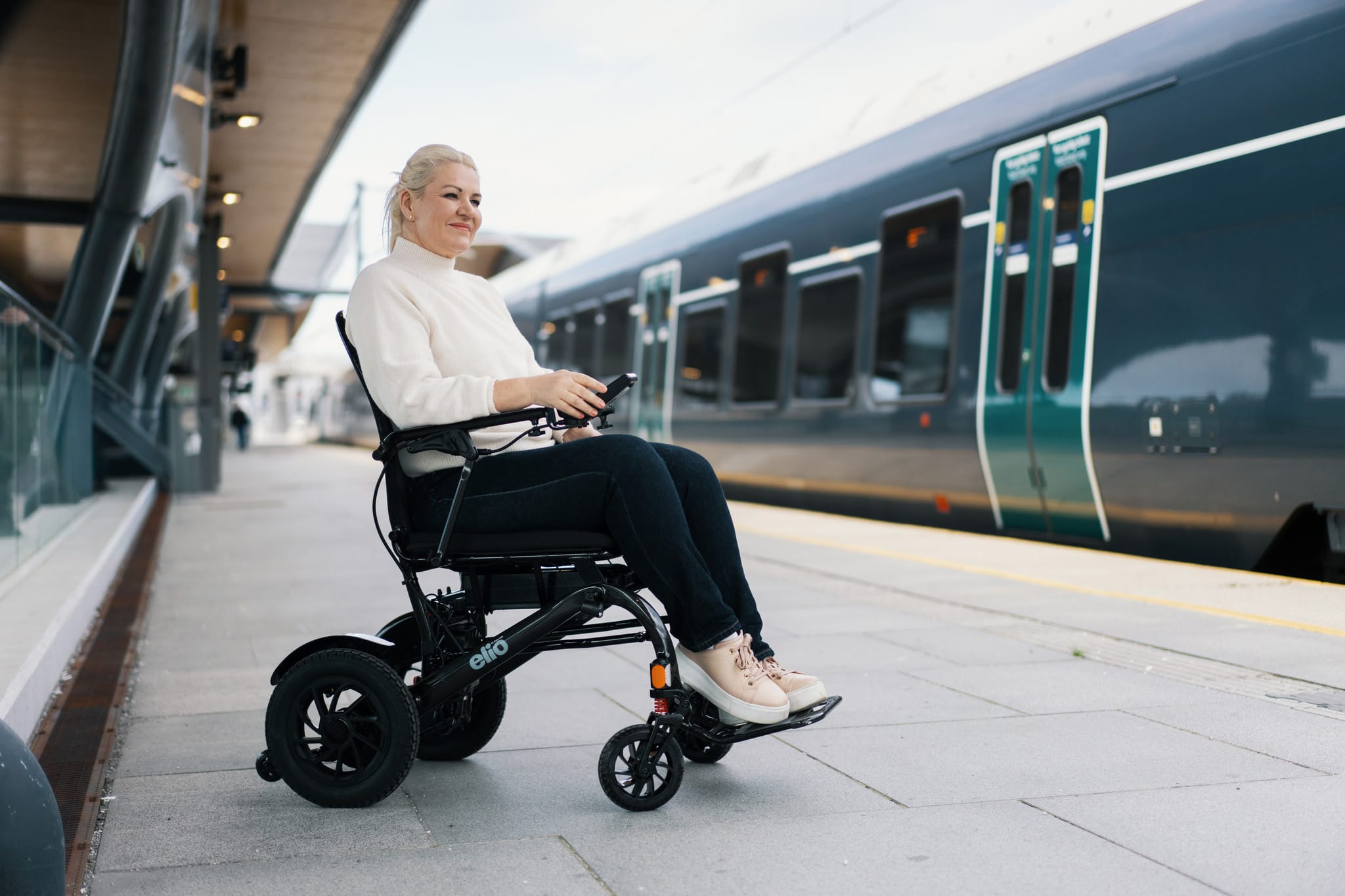 Marianne sitter i Elio elektrisk rullestol og venter på toget. Hun er iført hvit genser og mørk bukse. Toget står på perrongen.