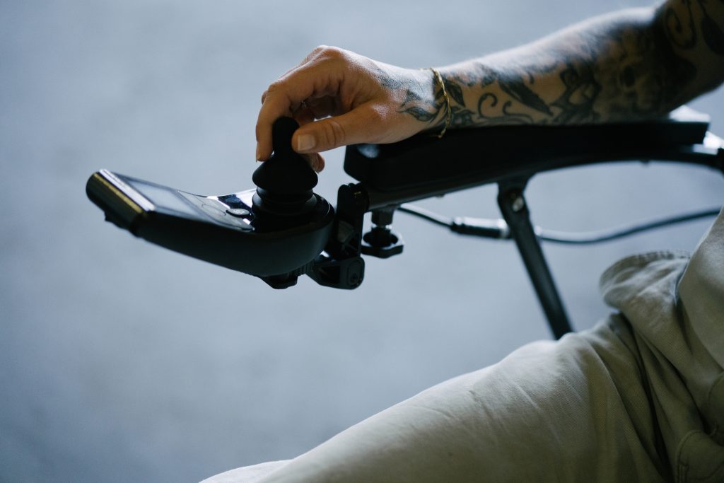 Nærbilde av joystick og en arm som hviler på armlenet og kjører Elio sammenleggbare elektriske rullestol. 