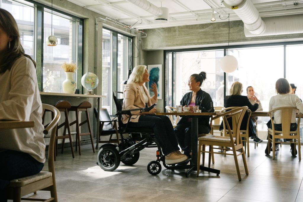 To venninner sitter rundt et bord på en café. Damen til venstre er iført pent tøy og sitter i elio sammenleggbare elektriske rullestol. Rundt dem sitter andre mennesker på cafeen. 