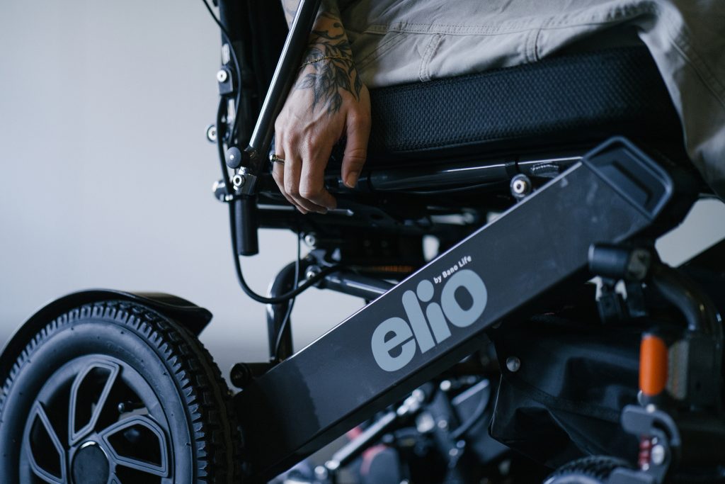 En person sitter i Elio sammenleggbare elektriske rullestol. Hånden holder i en knapp på siden av stolen på vei til å legge ned ryggen på stolen. 