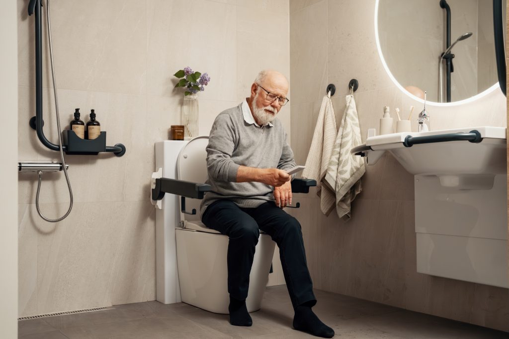 Bruker ser på fjernkontrollen, mens han sitter på Clean Comfort gulvstående toalett med spyl- og tørkefunksjoner. Toalettet er plassert på et moderne bad. 