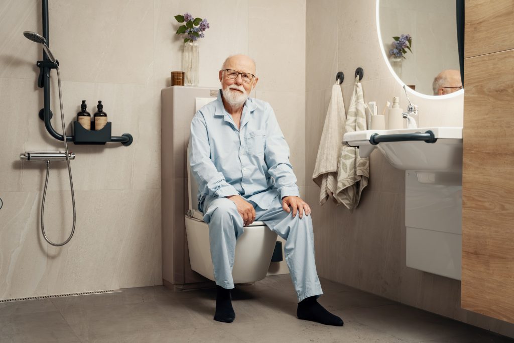 Stilig mann i pysj sitter på Clean Comfort vegghengt spyl- og tørketoalett. Smiler til kamera. På badet er det også en vask med støttehåndtak. Toalettet er montert rett ved dusjen.